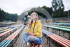 Young blond woman, wearing yellow hoody, blue jeans and eyeglasses, sitting on colorful bench in city urban park in summer.