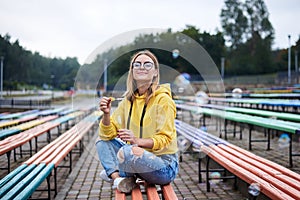 Young blond woman, wearing yellow hoody, blue jeans and eyeglasses, sitting on colorful bench in city urban park in summer.