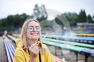 Young blond woman, wearing yellow hoody, blue jeans and eyeglasses, sitting on colorful bench in city urban park in summer. Close-
