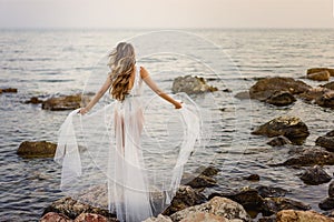 Young blond woman in white summer dress standing on the rocks and looking at the sea. Caucasian girl enjoys beautiful view at sunr