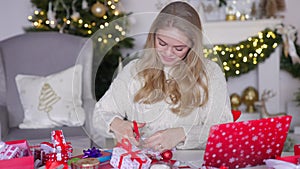 Young blond woman wrapping xmas gift box sitting next to table in living room