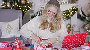 Young blond woman wrapping xmas gift box sitting next to table in living room