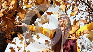 Young blond woman in yellow raincoat throwing autumn leaves happily in park