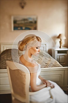 Young blonde beautiful woman with a perfect body and with stylish wedding hairstyle in a white dress sits in an armchair