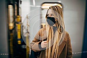 Young blonde in black medical mask looking at side while standing in subway near carriage