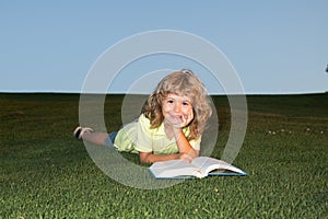 A young blonde boy is lying on the green grass outdoors reading a white book in summer. Smart child reading book, laying