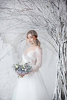 A young blonde bride in white wedding dress on a background of white walls and white tree in the background