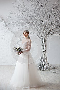 A young blonde bride in white wedding dress on a background of white walls and white tree in the background