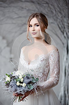 A young blonde bride in white wedding dress on a background of white walls and white tree in the background