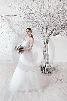 A young blonde bride in white wedding dress on a background of white walls and white tree in the background