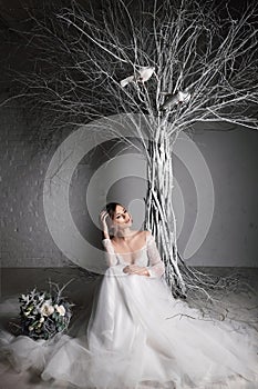 A young blonde bride in white wedding dress on a background of white walls and white tree in the background