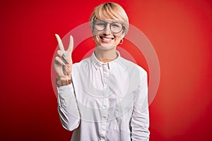 Young blonde business woman with short hair wearing glasses over red background showing and pointing up with fingers number two
