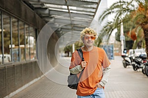 Young blonde caucasian guy dressed in stylish glasses with backpack keeps hand in his pocket.