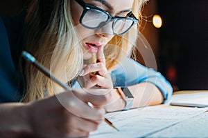 Young blonde caucasian student in eyeglasses thinking and writing something with pencil