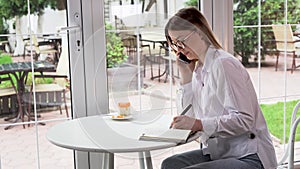 A young blonde Caucasian woman sits at a table in a cafÃ© communicating with someone on her smartphone makes notes in her