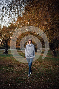 Young blonde Caucasian woman walking straight ahead looking at camera smiling wearing grey sweater, blue jeans and