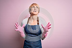 Young blonde cleaner woman with short hair wearing apron and gloves over pink background crazy and mad shouting and yelling with