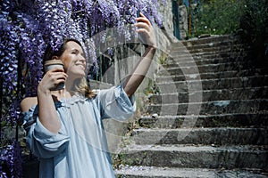 Young blonde curly hair woman in blooming wisteria garden in spring. Woman wearing in lilac dress making selfie on smartphone