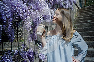 Young blonde curly hair woman in blooming wisteria garden in spring. Woman wearing in lilac dress walking with coffee