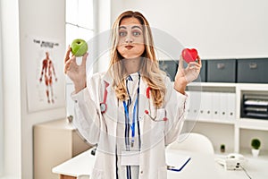 Young blonde doctor woman holding heart and green apple puffing cheeks with funny face