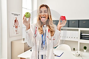 Young blonde doctor woman holding heart and green apple sticking tongue out happy with funny expression