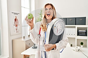 Young blonde doctor woman holding weighing machine and green apple angry and mad screaming frustrated and furious, shouting with