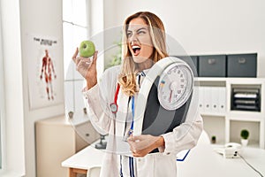 Young blonde doctor woman holding weighing machine and green apple angry and mad screaming frustrated and furious, shouting with