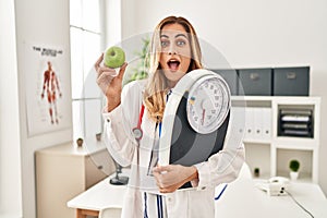 Young blonde doctor woman holding weighing machine and green apple celebrating crazy and amazed for success with open eyes