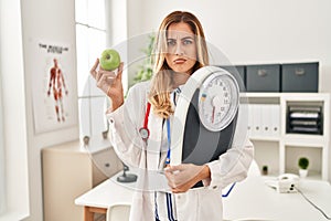 Young blonde doctor woman holding weighing machine and green apple depressed and worry for distress, crying angry and afraid