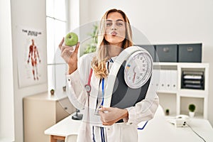 Young blonde doctor woman holding weighing machine and green apple looking at the camera blowing a kiss being lovely and sexy