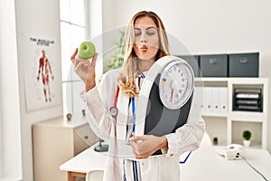Young blonde doctor woman holding weighing machine and green apple making fish face with mouth and squinting eyes, crazy and