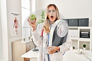 Young blonde doctor woman holding weighing machine and green apple in shock face, looking skeptical and sarcastic, surprised with
