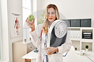 Young blonde doctor woman holding weighing machine and green apple skeptic and nervous, frowning upset because of problem