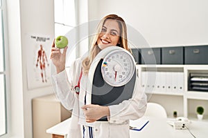 Young blonde doctor woman holding weighing machine and green apple smiling with a happy and cool smile on face