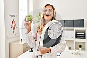 Young blonde doctor woman holding weighing machine and green apple smiling and laughing hard out loud because funny crazy joke