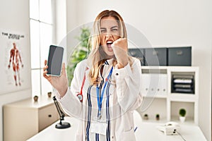 Young blonde doctor woman working at the clinic showing smartphone screen annoyed and frustrated shouting with anger, yelling