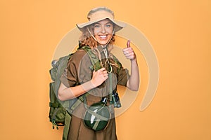 Young blonde explorer woman with blue eyes hiking wearing backpack and water canteen doing happy thumbs up gesture with hand