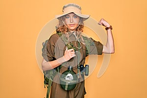 Young blonde explorer woman with blue eyes hiking wearing backpack and water canteen Strong person showing arm muscle, confident