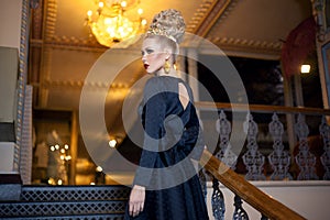 Young blonde female model, dressed in a long black dress with bow at the back, elegant hairstyle and crown and earrings.