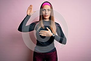 Young blonde fitness woman wearing sport workout clothes over isolated background Swearing with hand on chest and open palm,