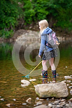 A young blonde girl with backpack and fishing net wades in the shallow water of a river