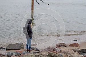 Young blonde girl in black jacket with hood looks into water, standing on gray stone among boulders on sandy shore of the sea