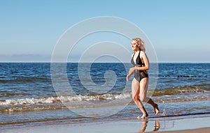 Young blonde girl in a black swimsuit runs along the sand of the sea shore
