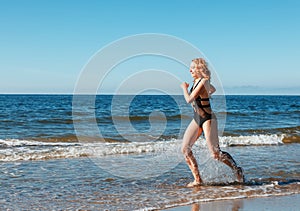 Young blonde girl in a black swimsuit runs along the sand of the sea shore