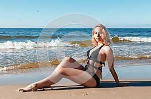 Young blonde girl in a black swimsuit sits on the sand of the sea shore