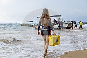 Young blonde girl in diving suit carries a medical case with first aid to the boat, which is pushed out by a group of