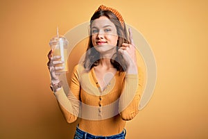 Young blonde girl drinking a glass of fresh healthy orange juice over yellow isolated background surprised with an idea or