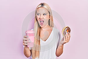 Young blonde girl eating doughnut and drinking coffee angry and mad screaming frustrated and furious, shouting with anger