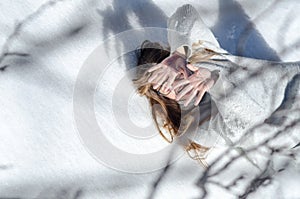 Young blonde girl with glasses lying on top of snow and covering her face with her hands