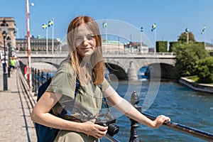 Young blonde girl in a green summer dress with a professional camera stands near the river in Stockholm on a sunny day